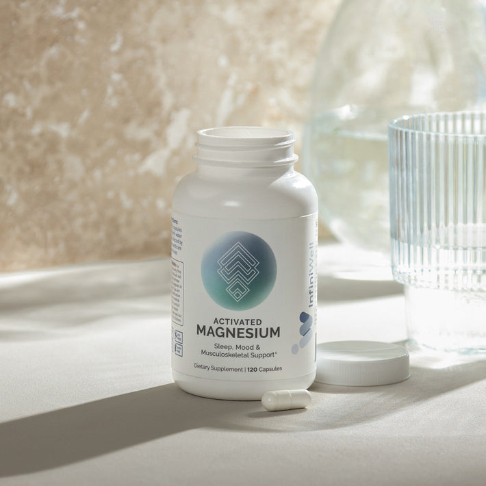 Infiniwell Activated Magnesium supplement bottle with 120 capsules, open cap and glass of water on light surface