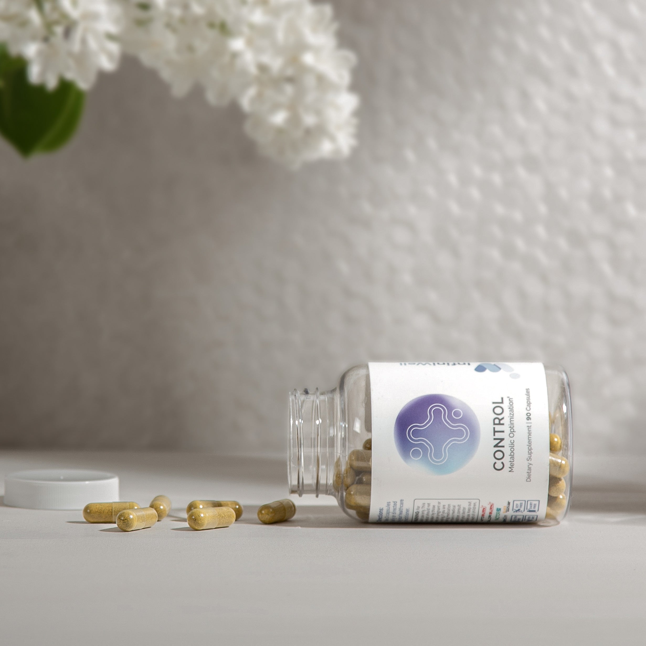 Olive-toned capsules spilled from a clear bottle, near white lilac blooms in soft-focus.