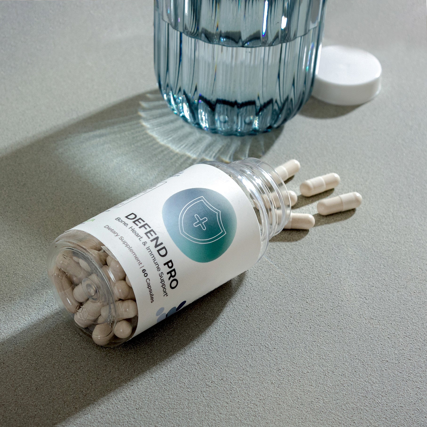 Beige capsules spilling from a clear bottle, next to a glass of water, on a gray surface.