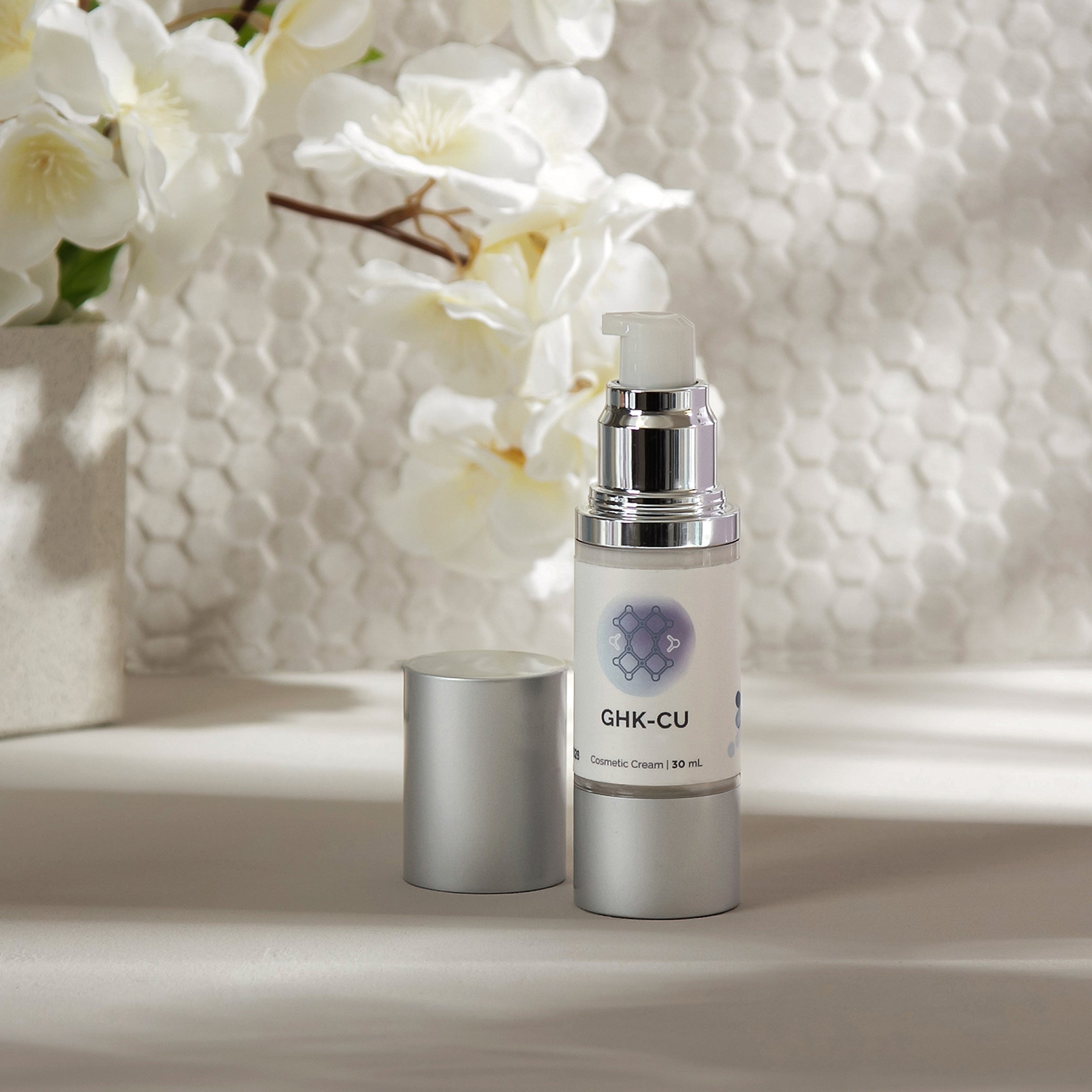 Silver cosmetic cream bottle with pump dispenser, 30ml, displayed on a light surface with white flowers.