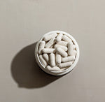 Top-down view of an open InfiniWell InfiniAGE supplement jar filled with white capsules on a neutral background.