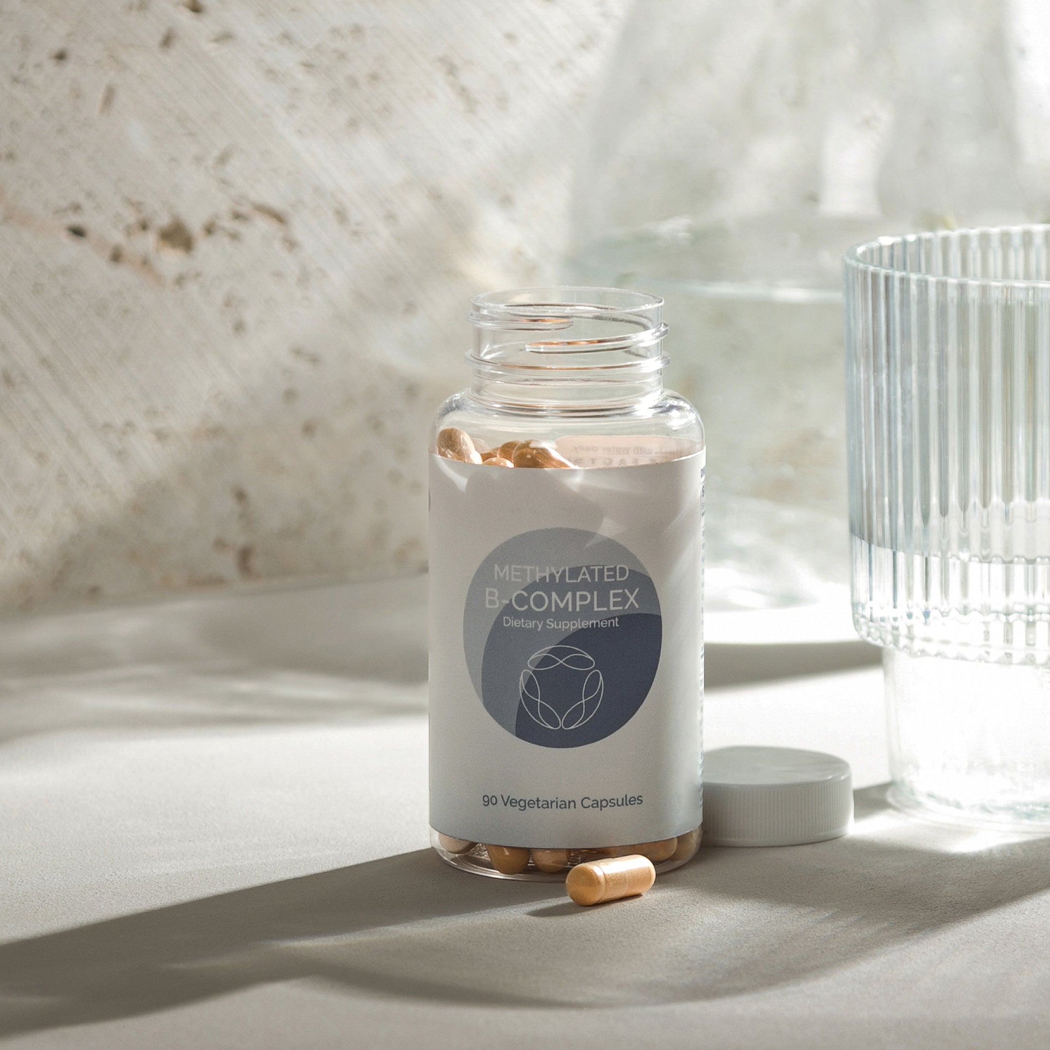 90 vegetarian capsules of B-complex dietary supplement in clear bottle, next to glass; light background.