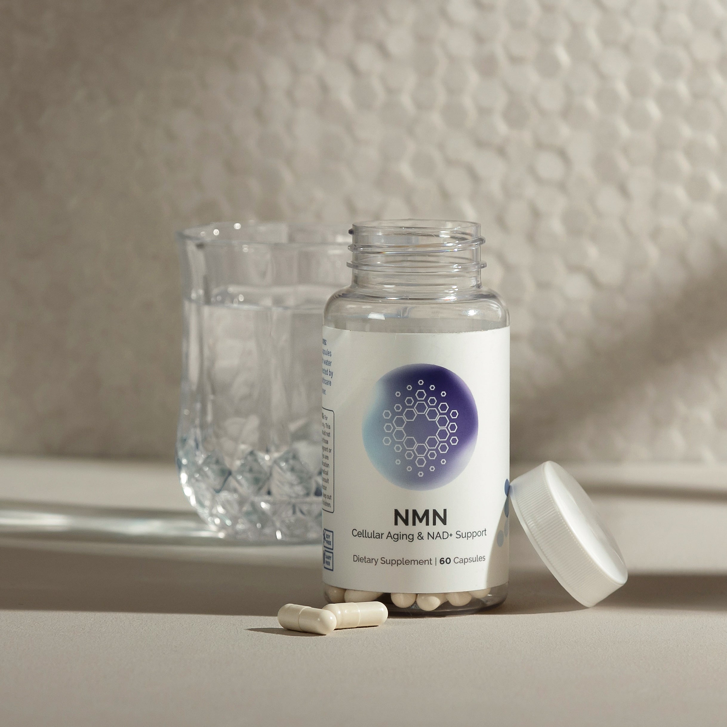 NMN supplement bottle, 60 capsules, with two capsules spilled nearby and a glass of water in the background.