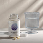 InfiniWell Senescent Regulator dietary supplement bottle with open lid, capsules, and glass of water on a neutral surface.