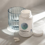 Infiniwell Activated Magnesium supplement bottle with open cap, two capsules and glass of water on light surface