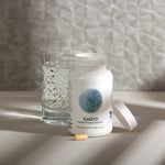 nfiniWell CoQ10 supplement bottle with open lid, capsule, and glass of water on a tabletop.
