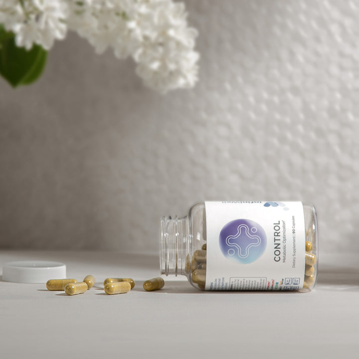 Olive-toned capsules spilled from a clear bottle, near white lilac blooms in soft-focus.

