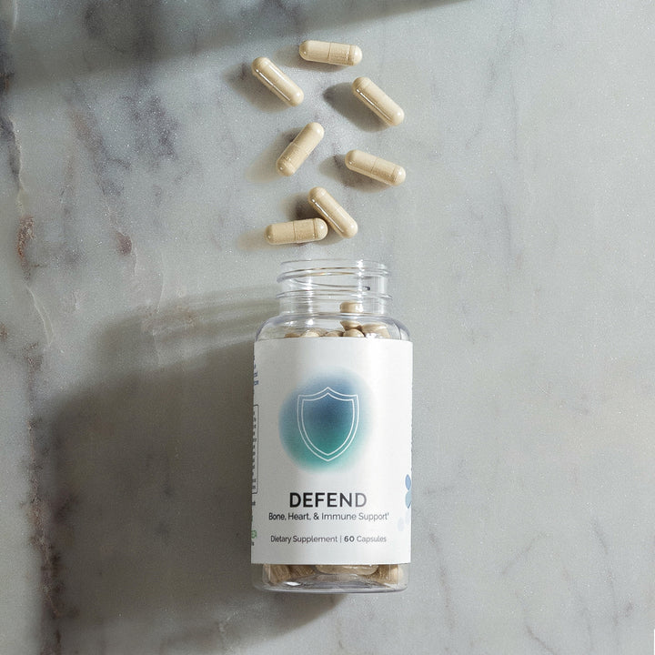 Beige capsules spilled from a clear bottle on a marble surface; 60 capsules total, supporting bone, heart & immune health.
