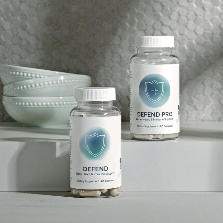 Two bottles of dietary supplement capsules promoting bone, heart, and immune support, displayed on a minimalist white surface.
