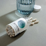 InfiniWell Defend Pro dietary supplement bottle lying on its side with spilled capsules beside a glass of water.