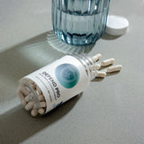 Beige capsules spilling from a clear bottle, next to a glass of water, on a gray surface.
