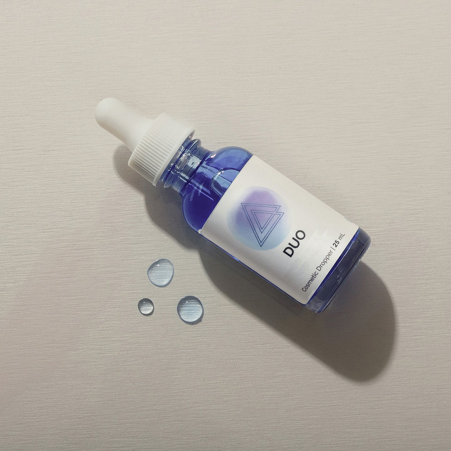 Infiniwell DUO cosmetic serum dropper bottle with blue liquid and water droplets on light textured surface, 25 mL