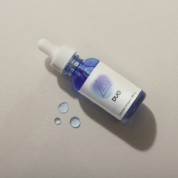 Infiniwell DUO cosmetic serum dropper bottle with blue liquid and water droplets on light textured surface, 25 mL