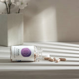 Dietary supplement capsules spilled from a clear bottle onto a white surface, bathed in soft sunlight.
