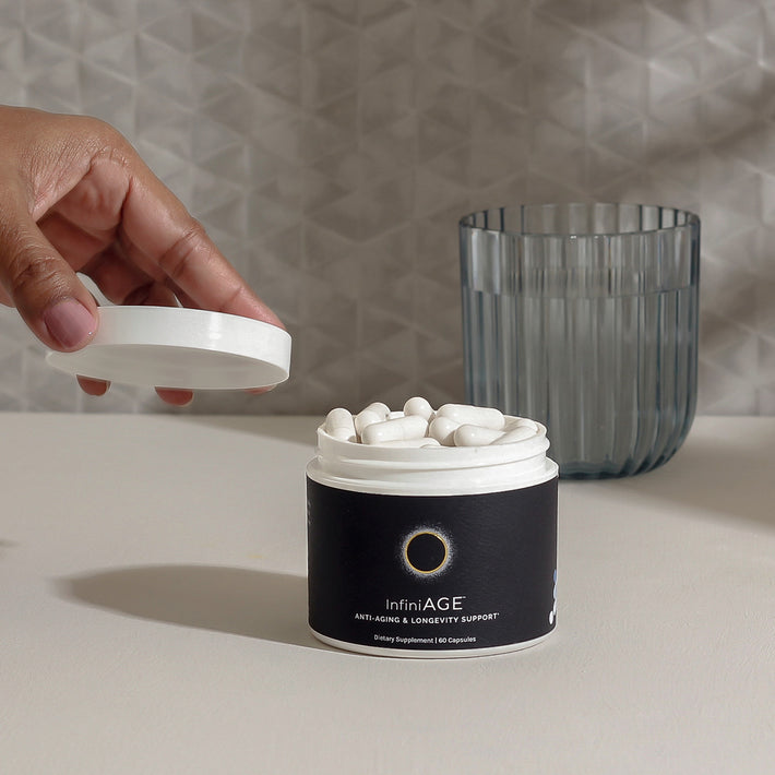 Open jar of InfiniAGE longevity supplement with white capsules beside a glass and hand lifting the lid.
