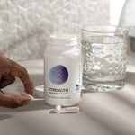 InfiniWell STRENGTH skeletal muscle support supplement bottle with 90 capsules beside a glass of water and loose capsules.