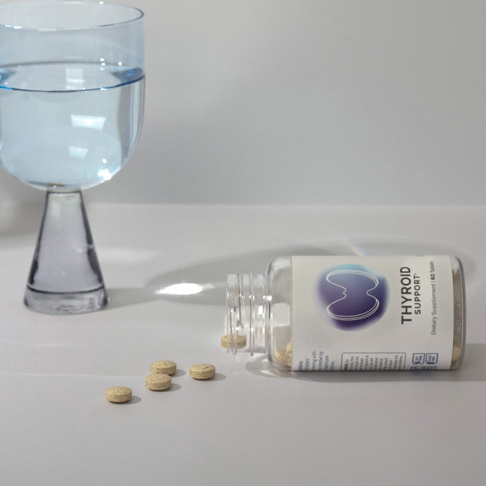 Infiniwell Thyroid Support supplement bottle on side with 60 tablets, spilled tablets and glass of water nearby