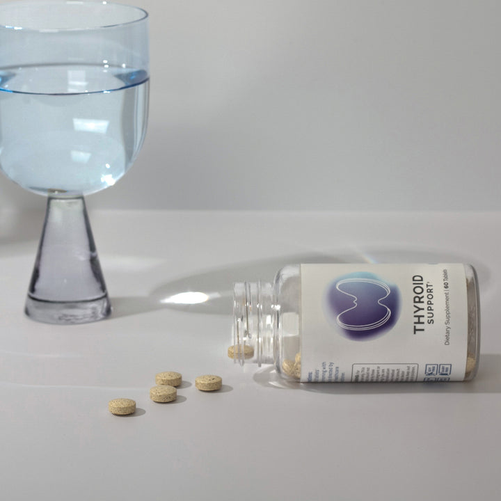 Beige tablets spilled from a clear supplement bottle, near a glass of water.
